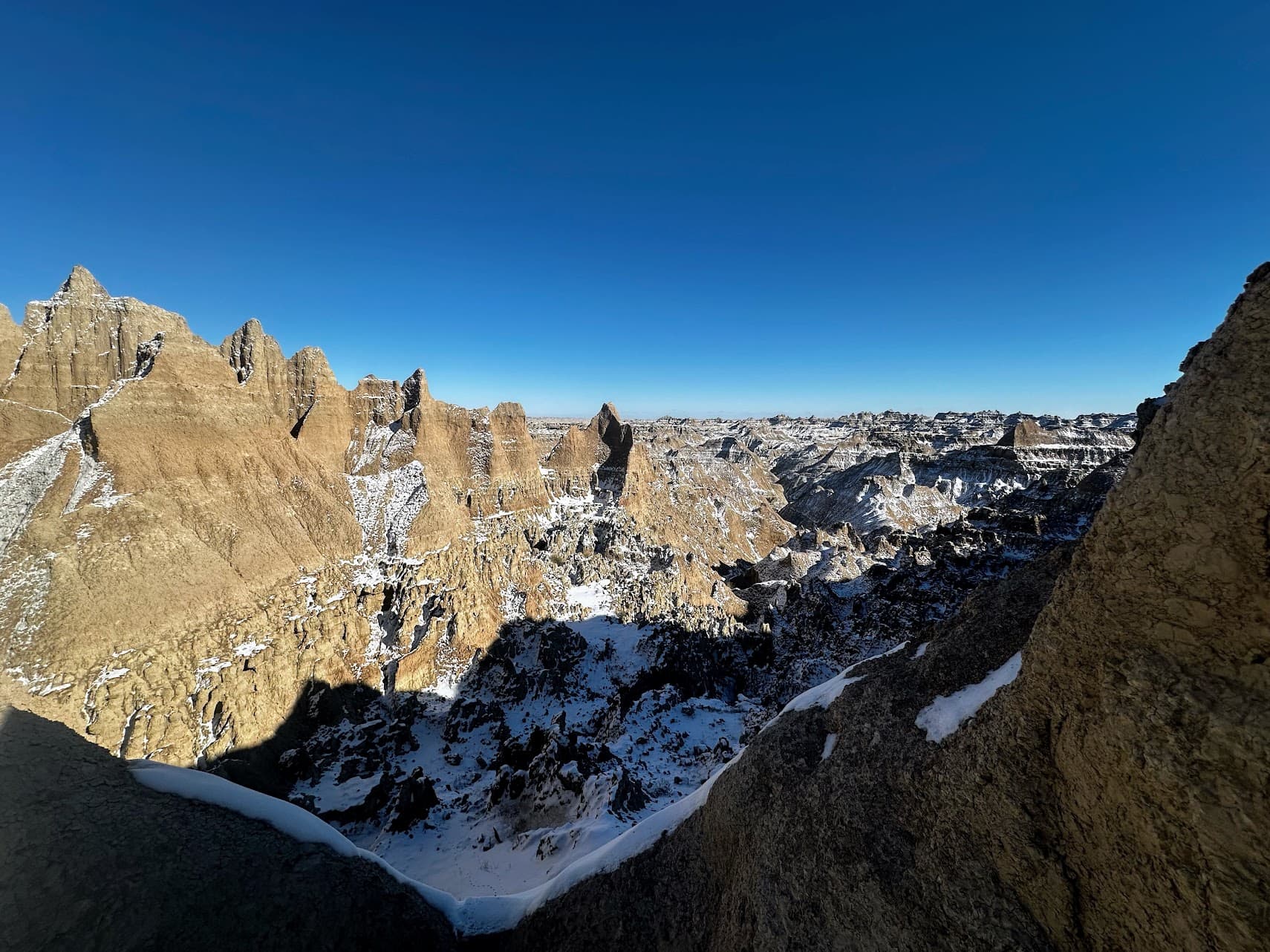 Sunlit jagged rock formations above a snow-covered ravine under a blue sky.